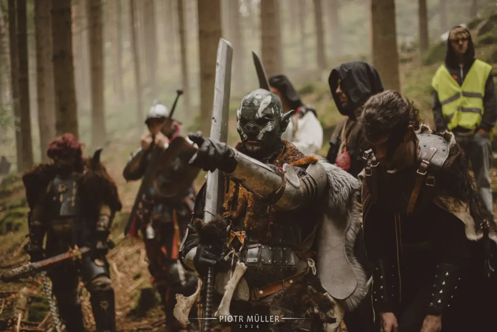 Ork w zbroi fantasy z mieczem, wskazujący ręką, w tle wojownicy – film LARP