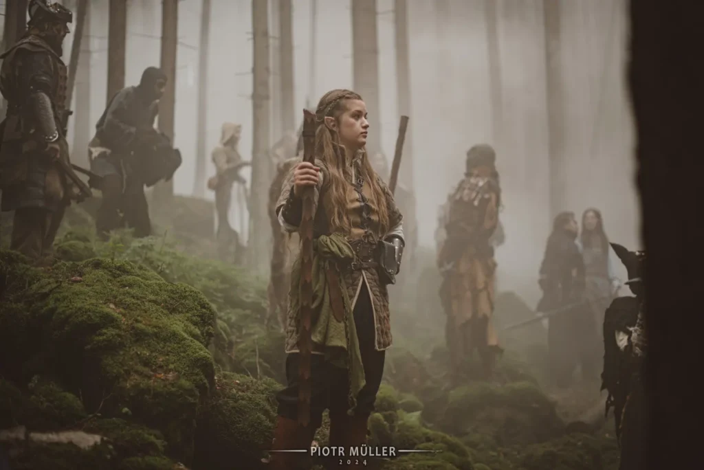 Młoda aktorka w stroju wojowniczki fantasy z kosturem, stojąca w lesie na planie filmu LARP